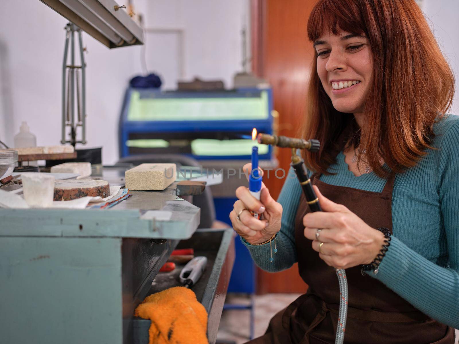 Front view of a a happy woman working in her artisan jewelry workshop sitting at a workbench lighting a torch to perform her work.