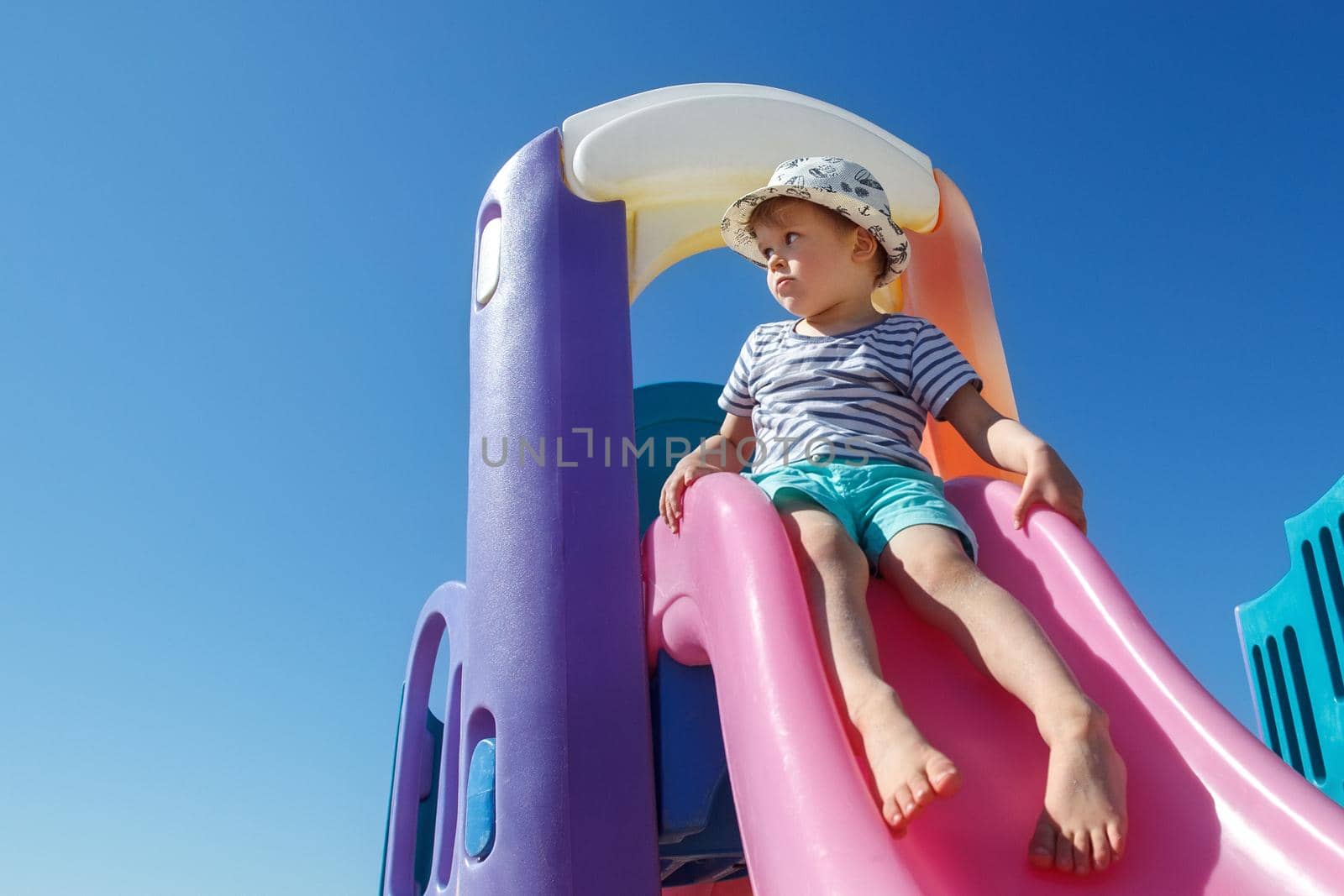 A little boy in a hat is ready to slide, on a colourful slide, he looks into the distance. There is free space for text in the background of the blue sky. by Lincikas