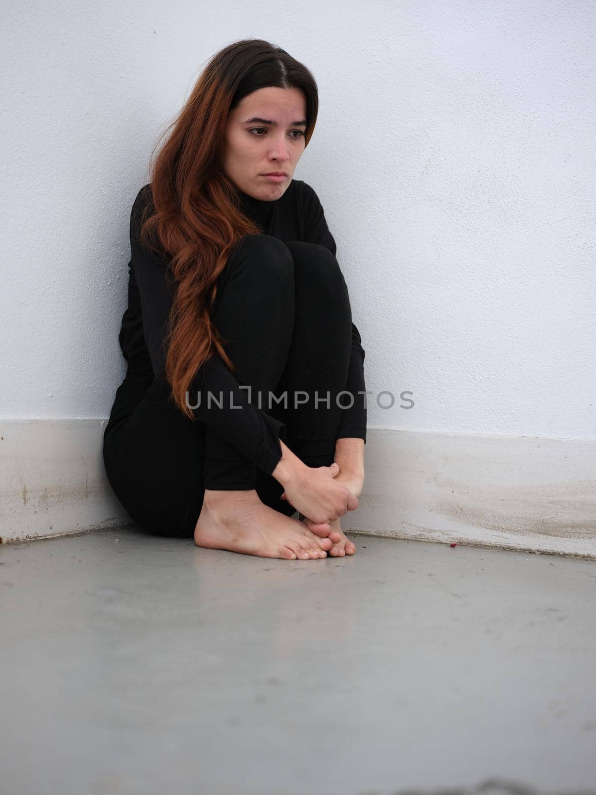 Vertical image of a young woman in the corner of a room dressed in black barefoot looking sad. Concept of emotions.