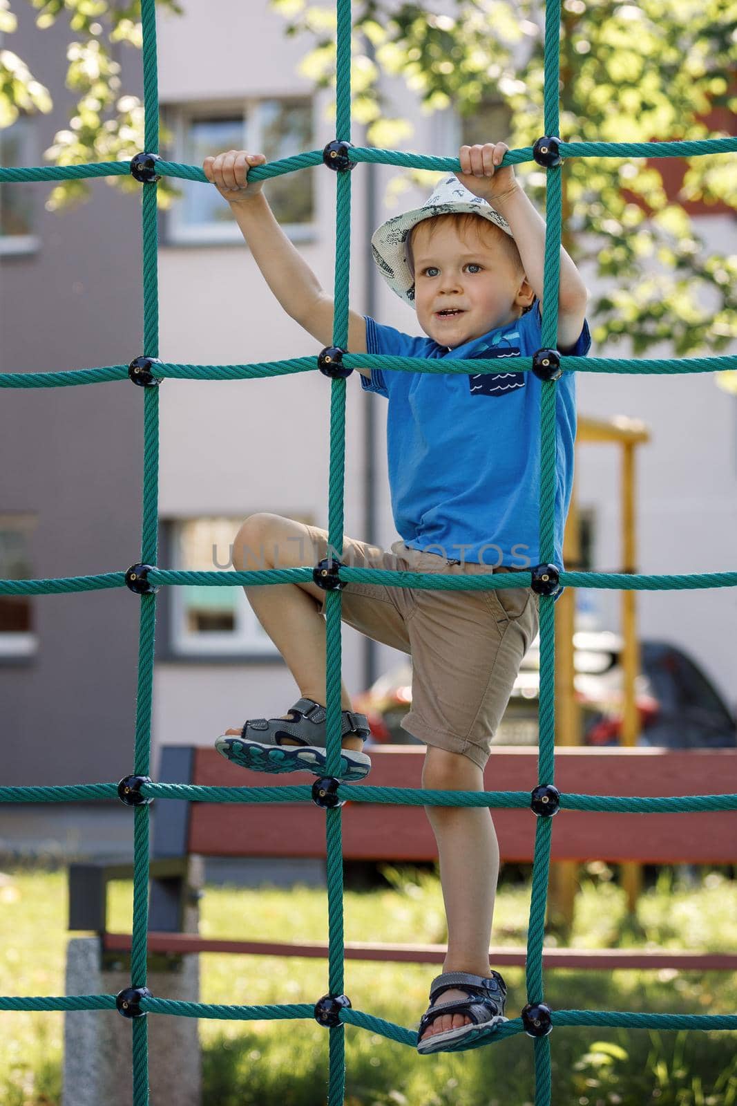 Child hanging on green rope net in city park. by Lincikas