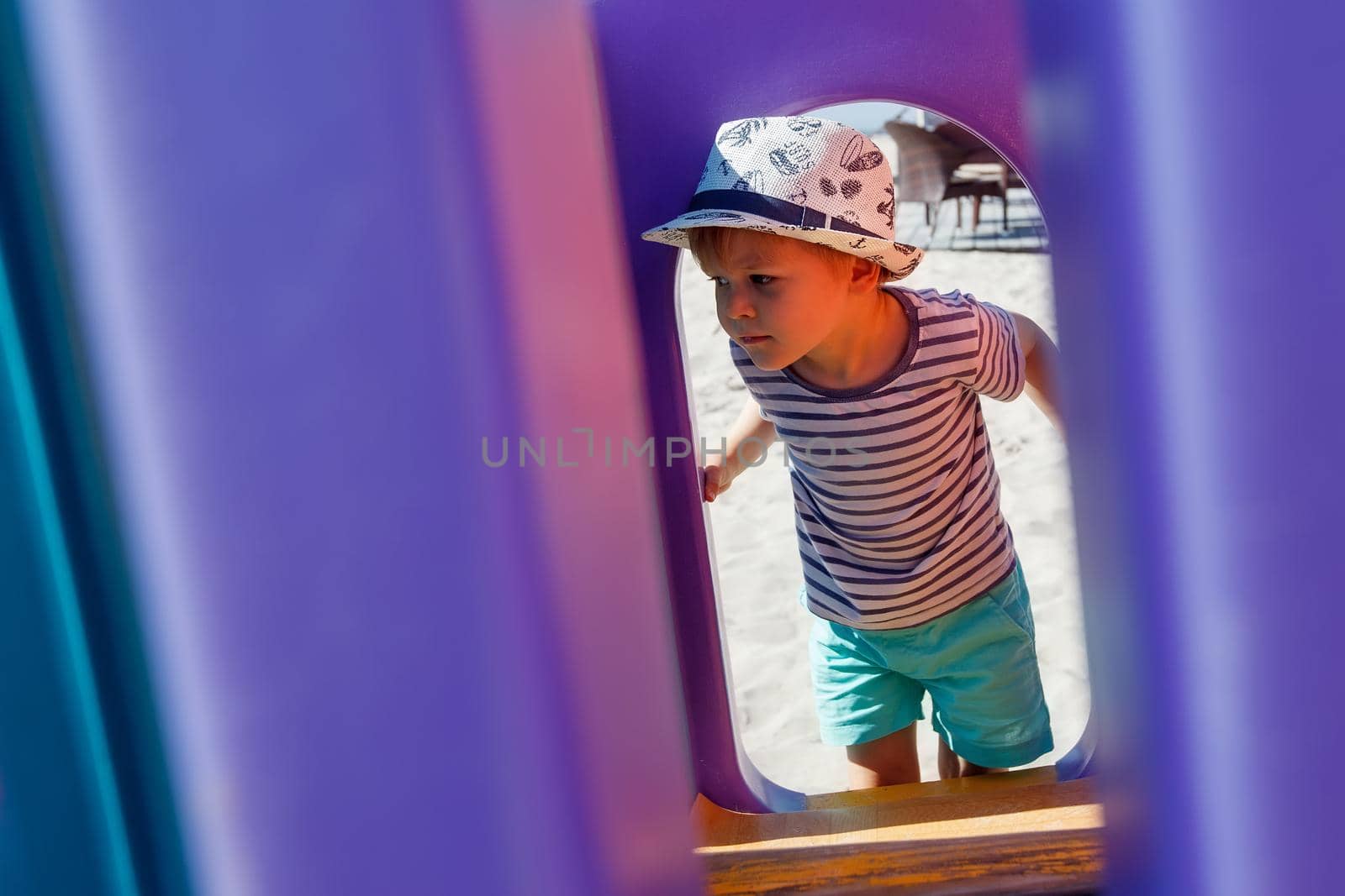 A little boy in a hat looks at the playground through a purple plastic arch. by Lincikas