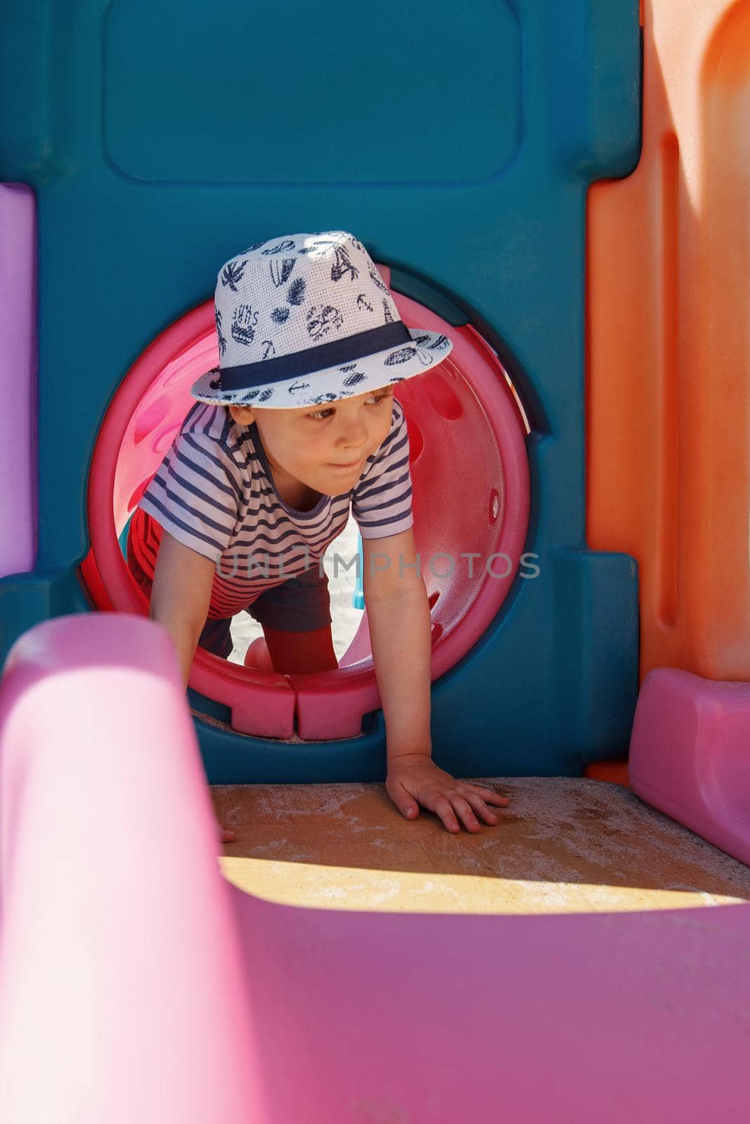 A little boy with a hat in a pink tunnel on a playground. The child is training, he is developing his physical skills