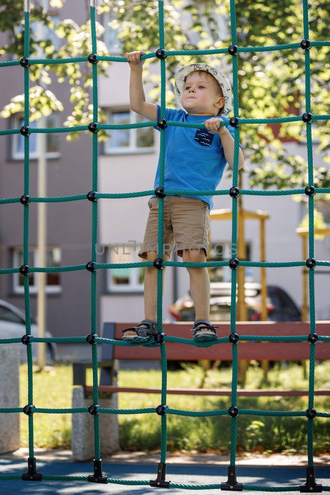 A little boy like a spider climbs up a green net of ropes on a playground.