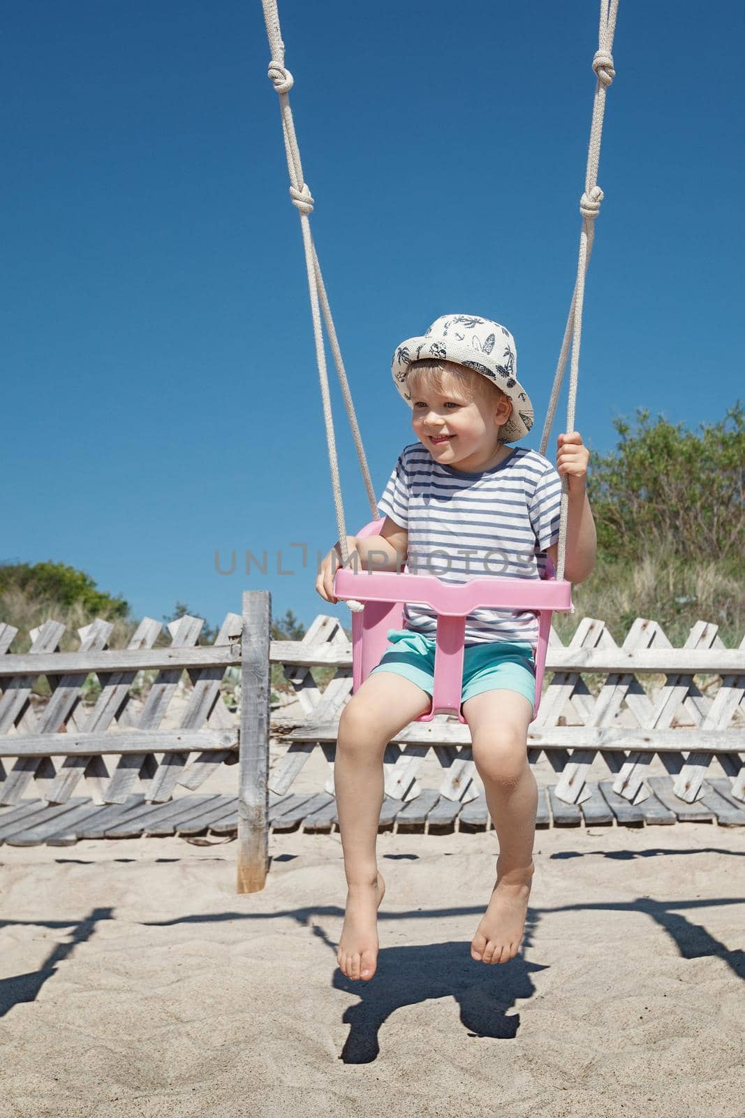 A cute boy swings on the beach. Pink plastic swing, blue sky and dune background. by Lincikas