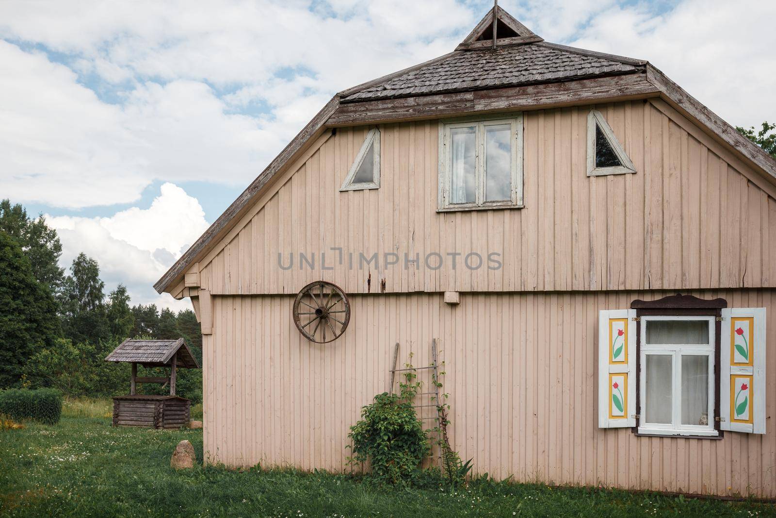 Old traditional wooden house with windows and shutters, rear view. An old carriage wheel is hung on the wall, the roof is covered with wooden tiles. by Lincikas