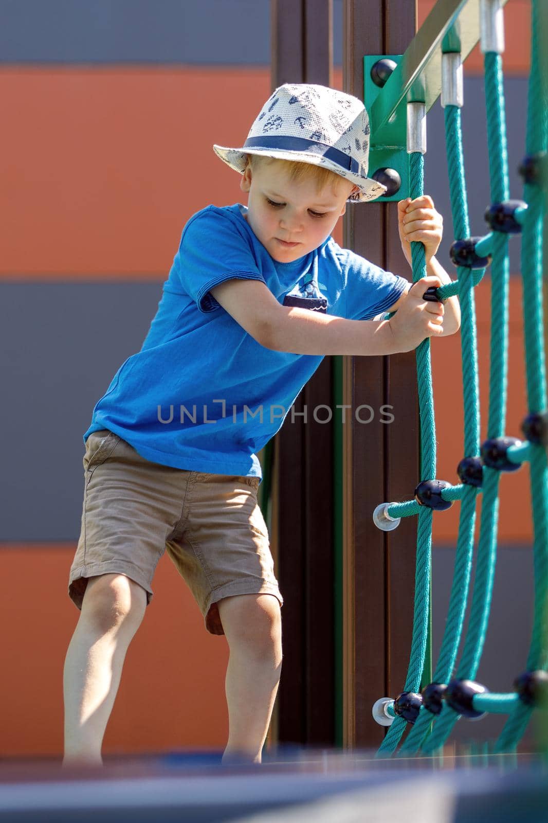 Low angle view of adorable little boy with hat having fun at playground. The child trains and carefully crosses the monkey bridge.