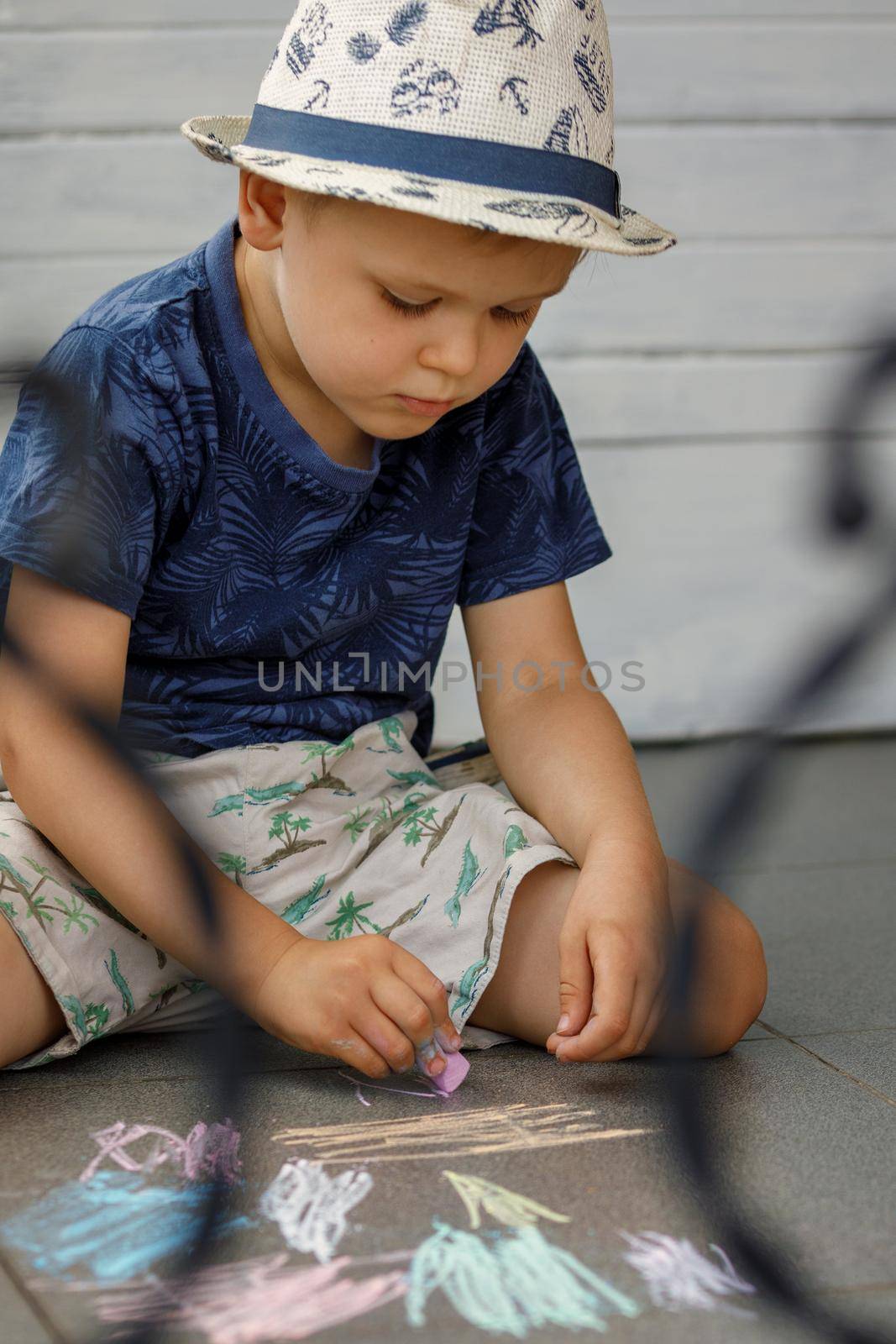 Little preschool boy draws with colorful chalks on the ground. The child is very focused on learning.