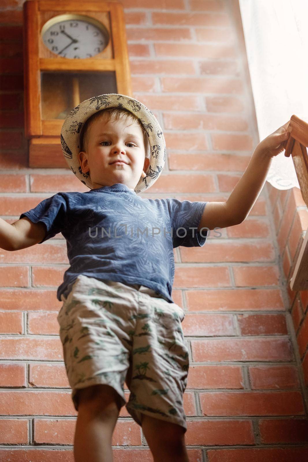 Portrait of a little boy from below, by the window in the sunlight, a red brick wall and an antique clock. by Lincikas