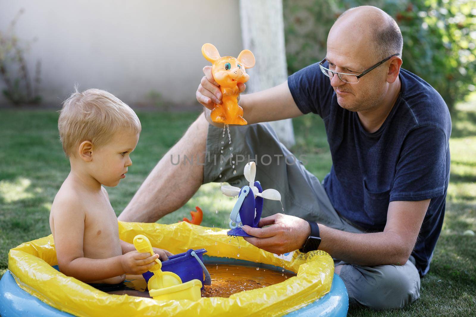 Little boy and middle age man sitting on a grass and play with water toys. Surprised looking at splashy toy. Family free time in outside.