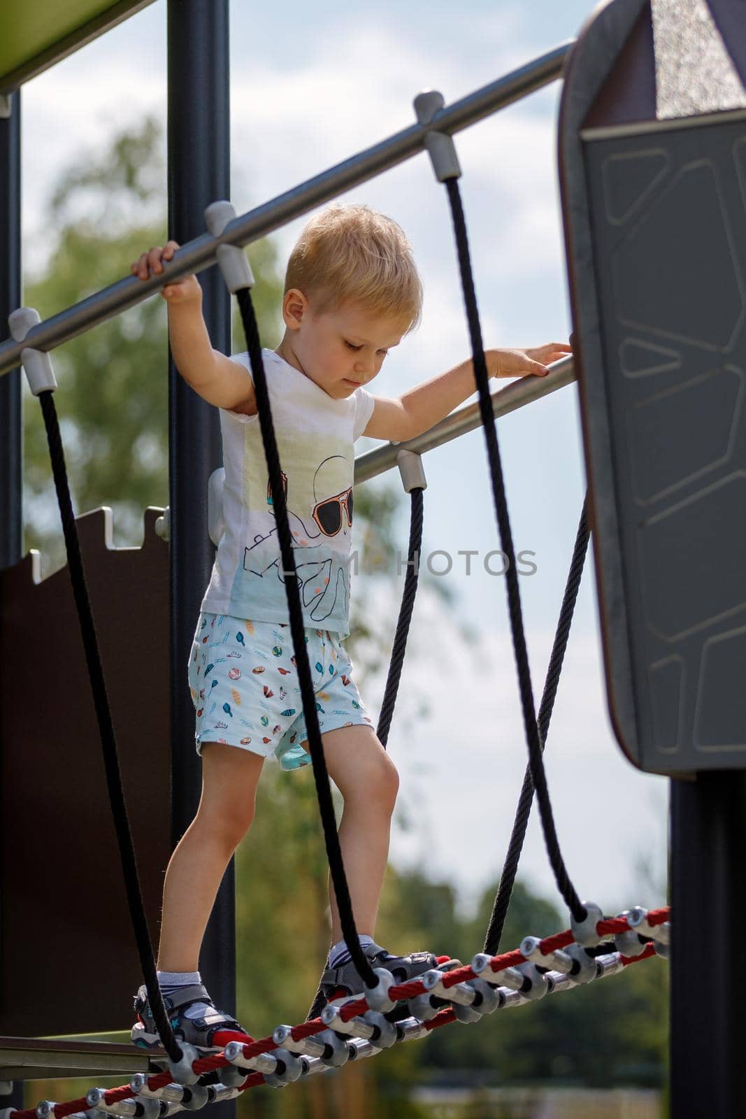 Low angle view of a little boy walking carefully across the playground monkey bridge by Lincikas