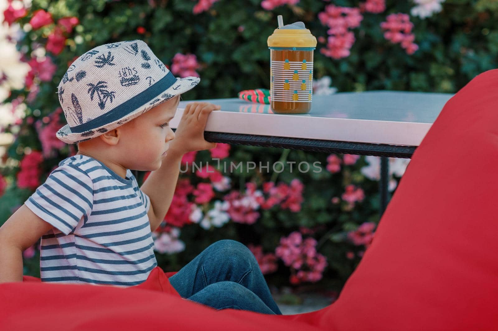 A little boy in a striped T-shirt is sitting at a table with juice sitting on a red armchair.