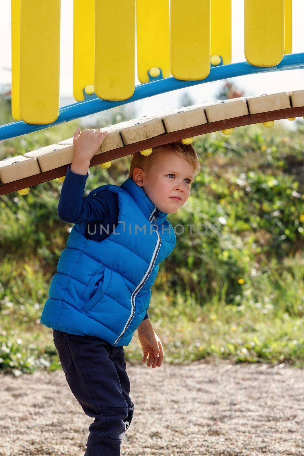 A little cute boy plays under a beautiful, curved bridge on a colorful playground. An agile child actively spends time in the fresh air. Vertical photo.