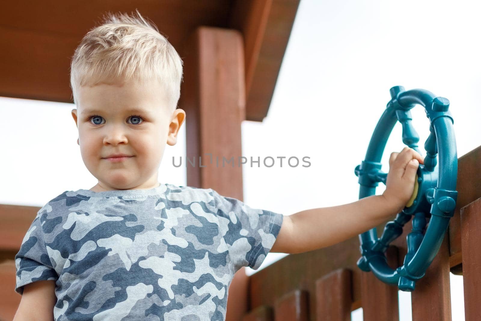 Pleased child is beeping a ship wheel in a playground. Kid wearing camouflage is making a signal  in a playhouse. Proudly beeping horn. Childhood full of adventure