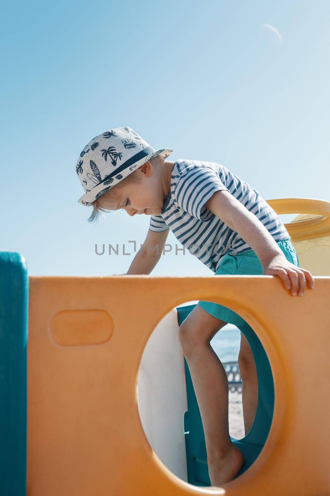 A little boy with a hat and a striped sailor shirt is playing on the beach playground, he is doing physical exercises