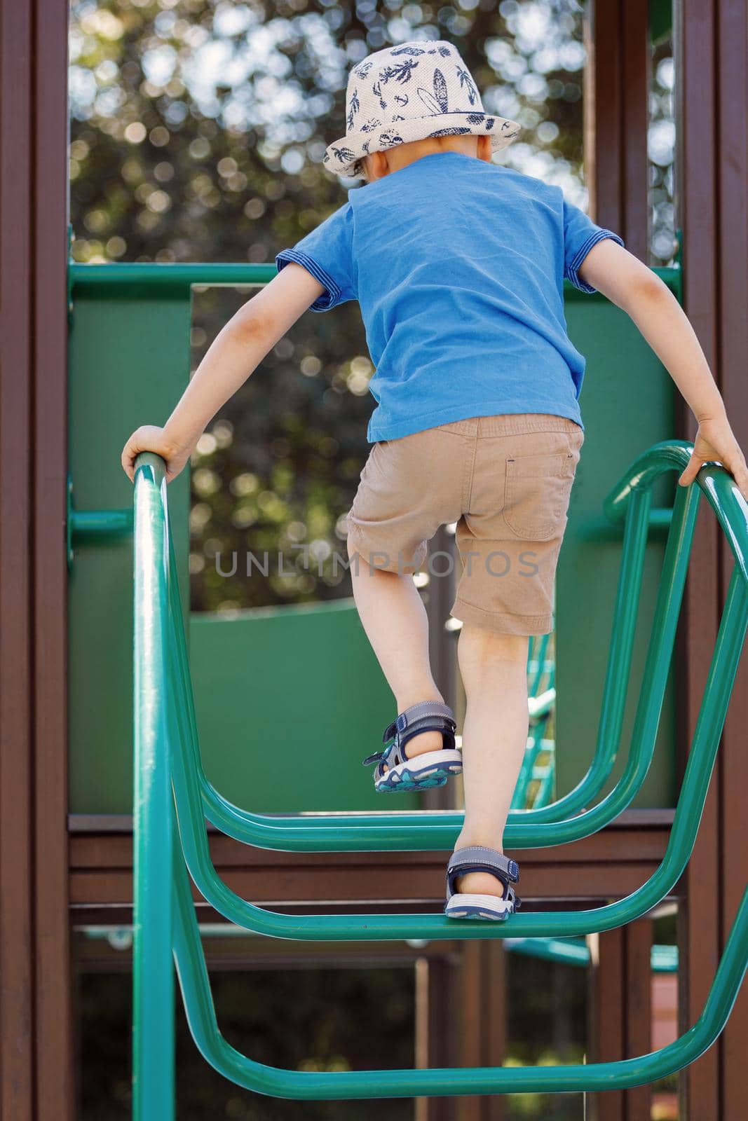 A little boy in a blue T-shirt boldly climbs the playground with a tubular green ladder, the child is not afraid to take risks. Rear view photo.