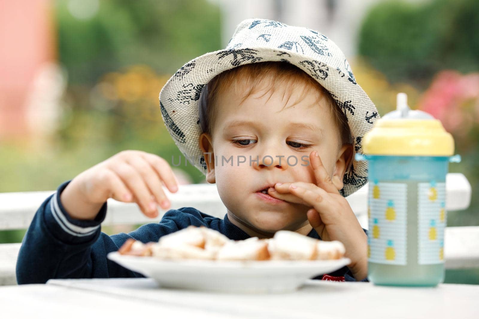 A little boy in a white hat tastes bread with cheese while licking his fingers. A plastic cup with pineapple juice on the table.