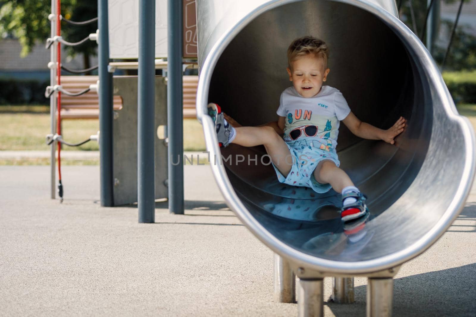Happy Kid Sliding Fast In Tube Slide On Playground Park. Joyful Boy Having Fun in Play Centre. Kindergarten For Elementary Age Children.