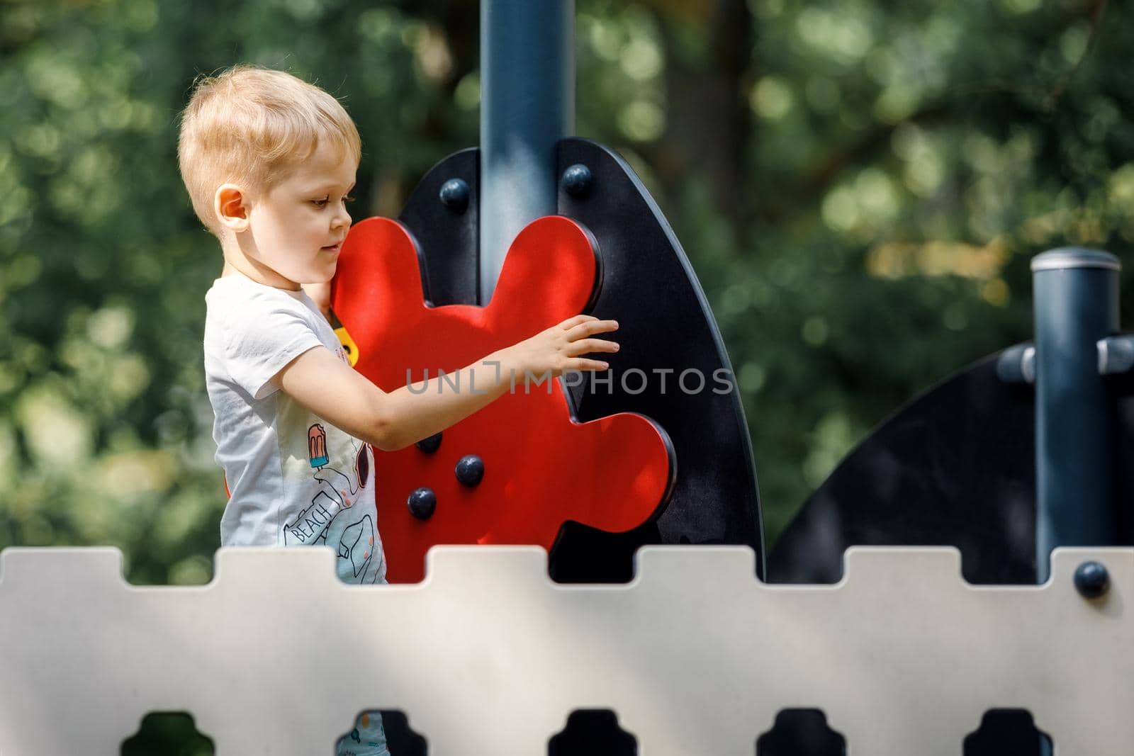 A cute boy on an outdoor playground, a red ship's helm, a decorative fence, a green foliage background.