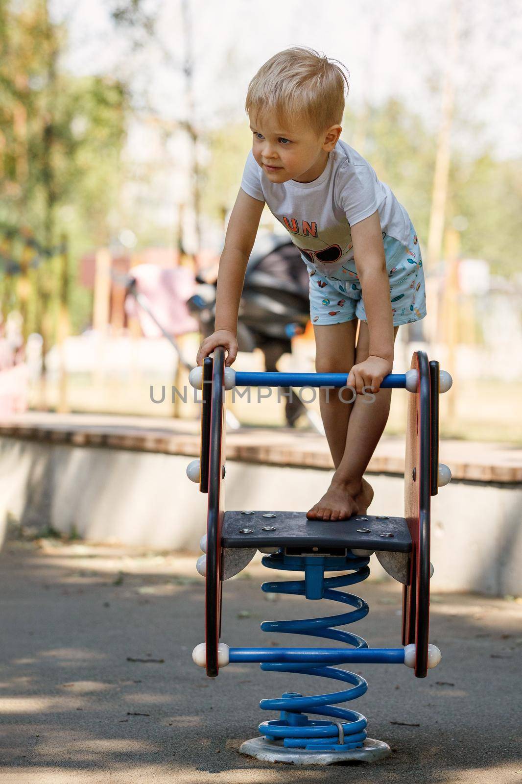 The brave little boy stands up and balances on a spring swing on the playground by Lincikas