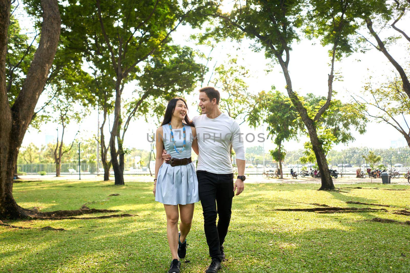 Multiethnic marriage walking while embracing looking each other in a park surrounded by trees