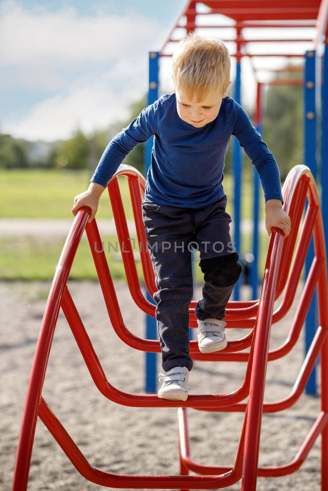 Brave small child playing and climbing arch ladder in public kid playground during beautiful day in summer time.