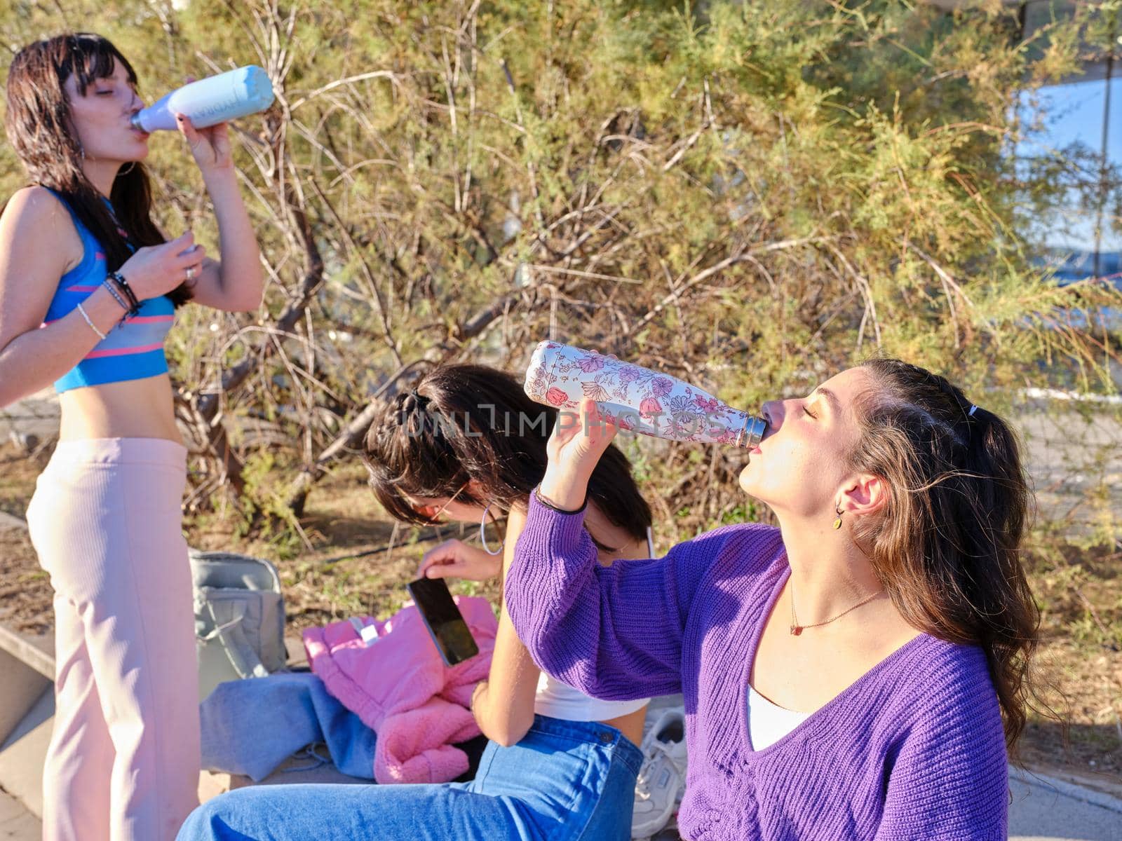 Three young women wearing vintage clothes drinking water after skate in a park during a sunny day