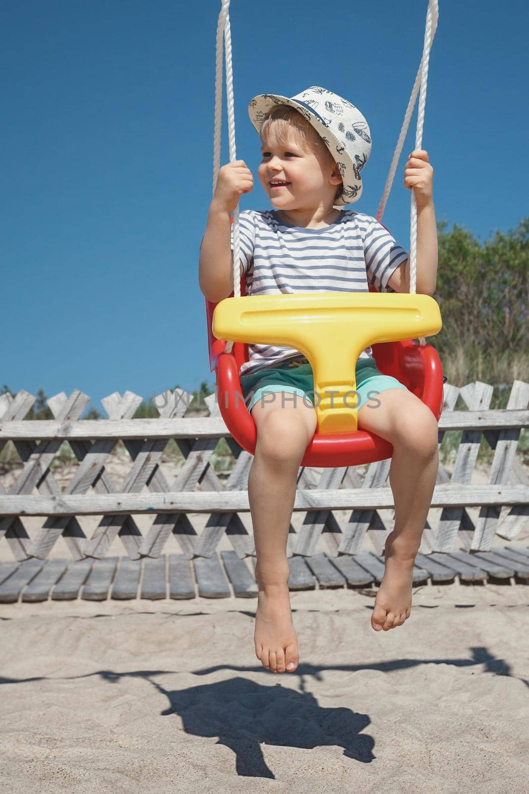 The beach, the blue sky and the happy boy swing in a red-yellow swing. The concept of weekend-rest, happy and carefree childhood. by Lincikas