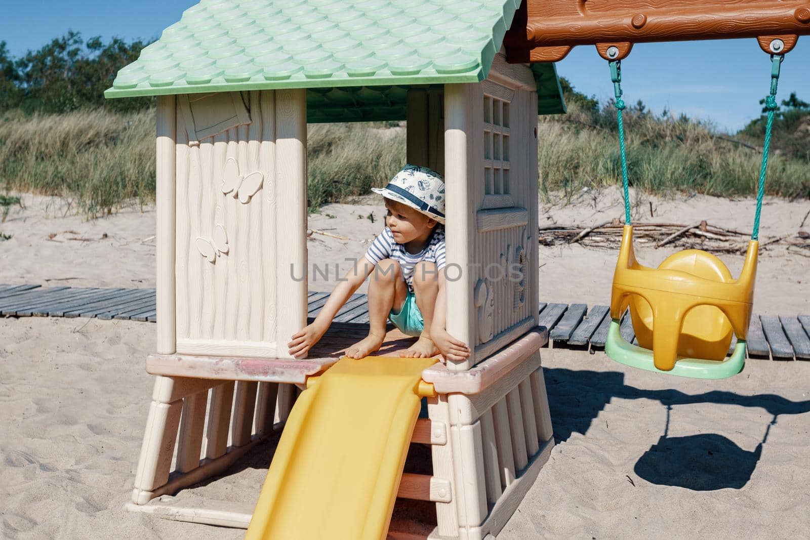 Children's playground on the beach, swings, slide, small hut and a cute child inside. by Lincikas