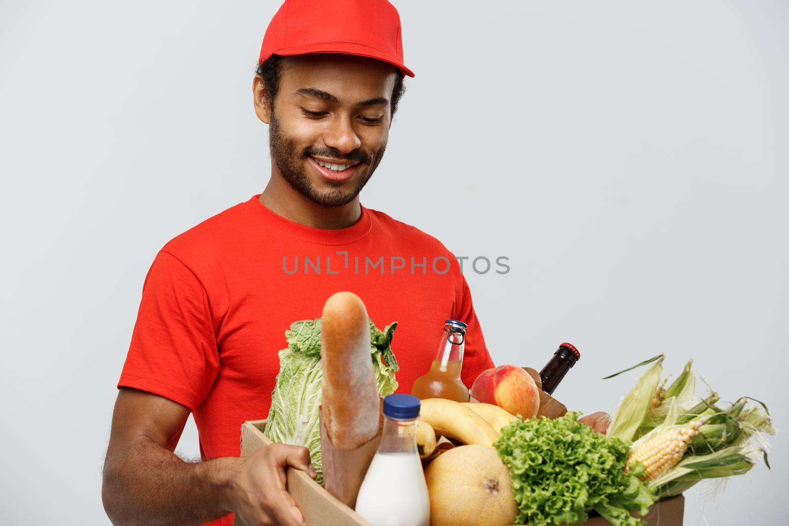 Delivery Concept - Handsome African American delivery man carrying package box of grocery food and drink from store. Isolated on Grey studio Background. Copy Space. by Benzoix