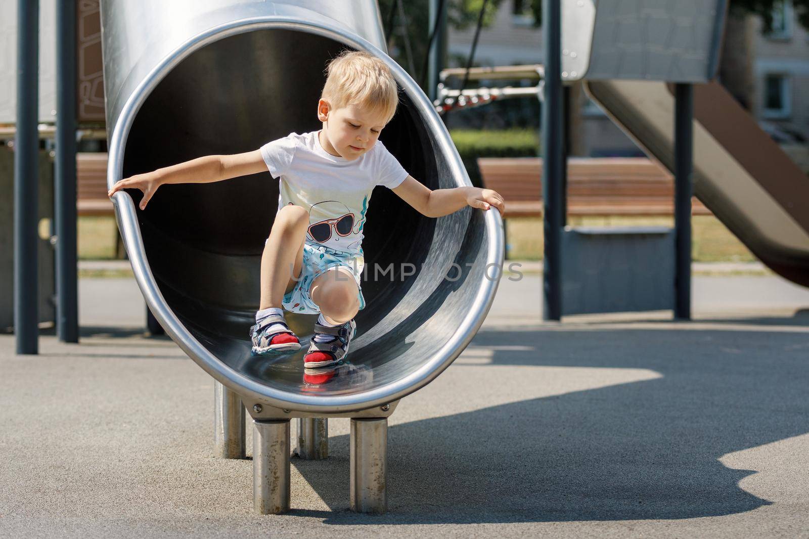 Boy child climbing sliding and playing on metal slide on outdoor playground in summer park. Activity and amusement center in kindergarten or school yard. Toddler kid outdoors.