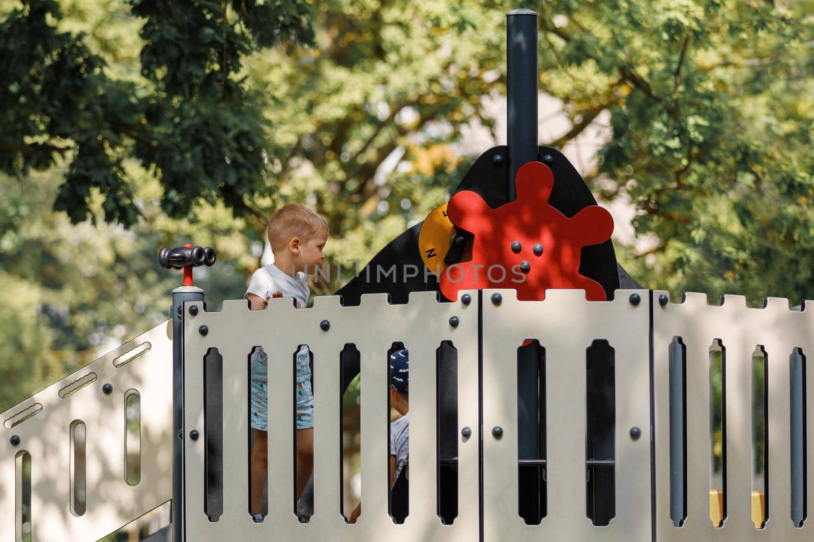 A cute boy on an outdoor playground, a red ship's helm, a decorative fence, a green foliage background. by Lincikas