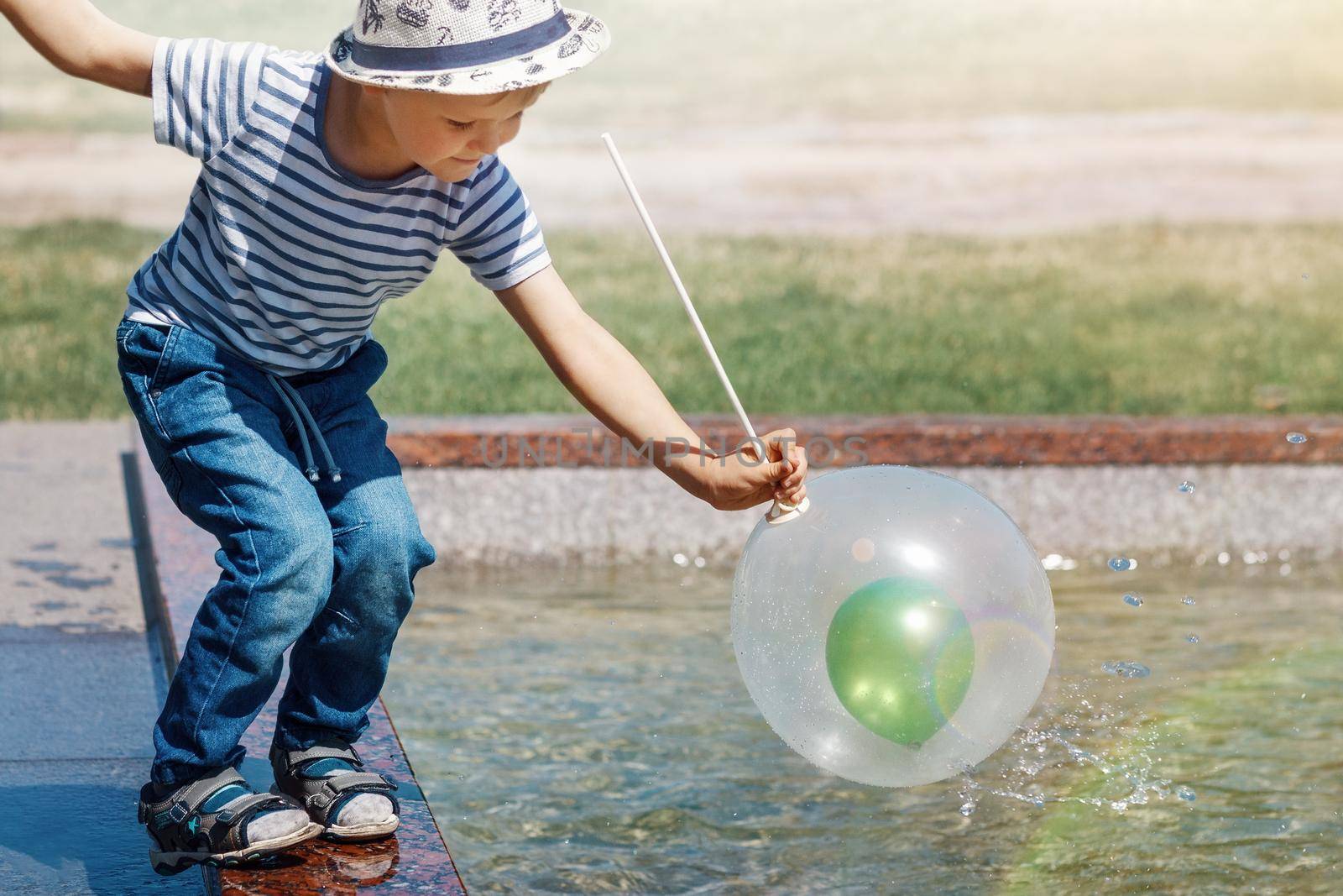 A little boy in a striped shirt and hat in the city by the fountain plays with white and green air balloons by Lincikas