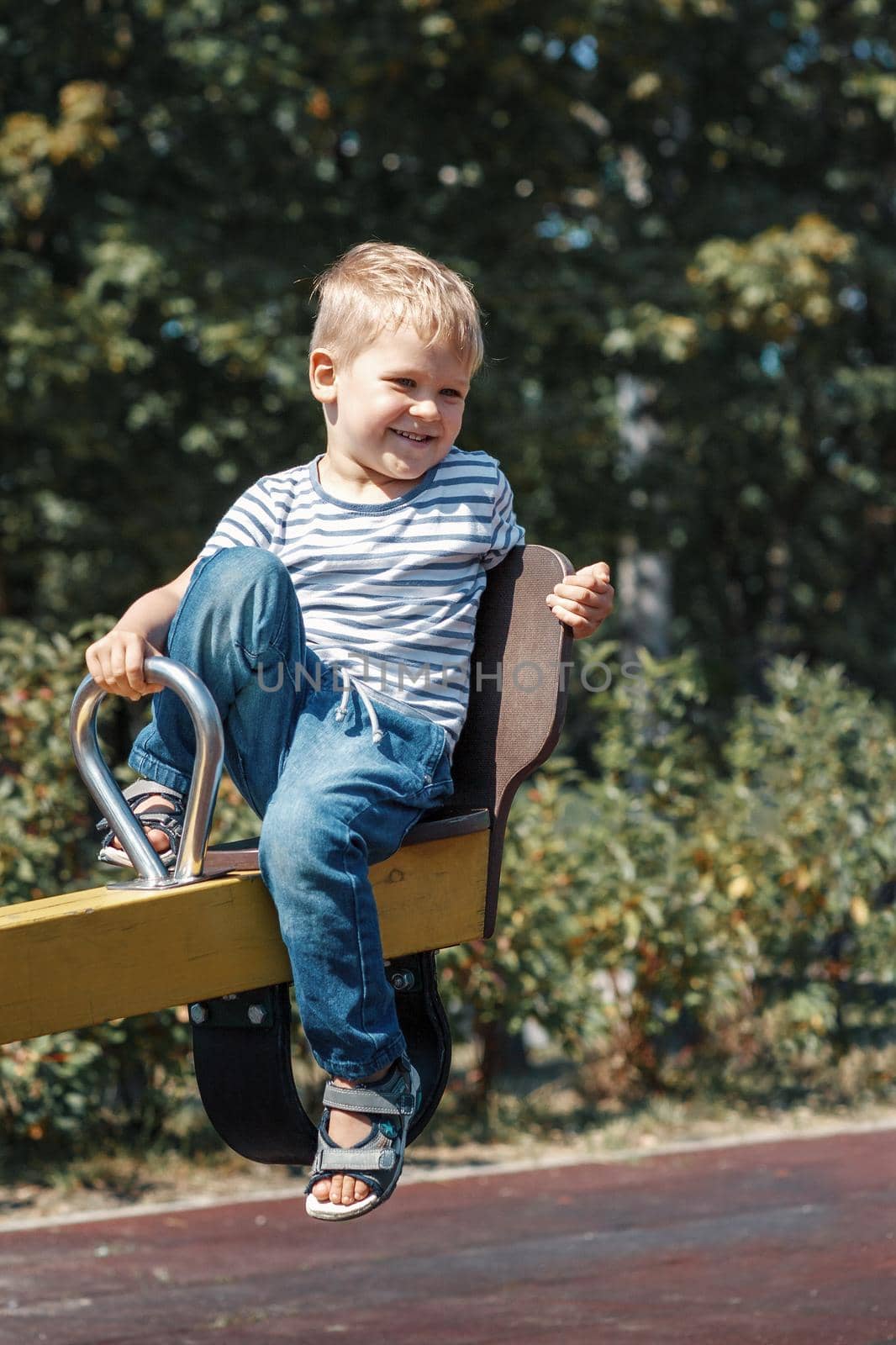 A cheerful, smiling, little boy on a swing on a city playground against a green foliage background.