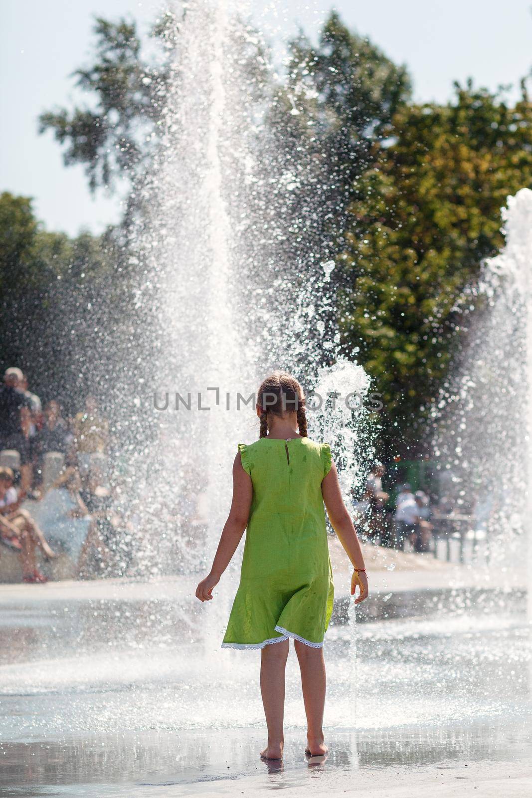A young girl in a light green dress barefoot dances by a fountain in the city center. by Lincikas
