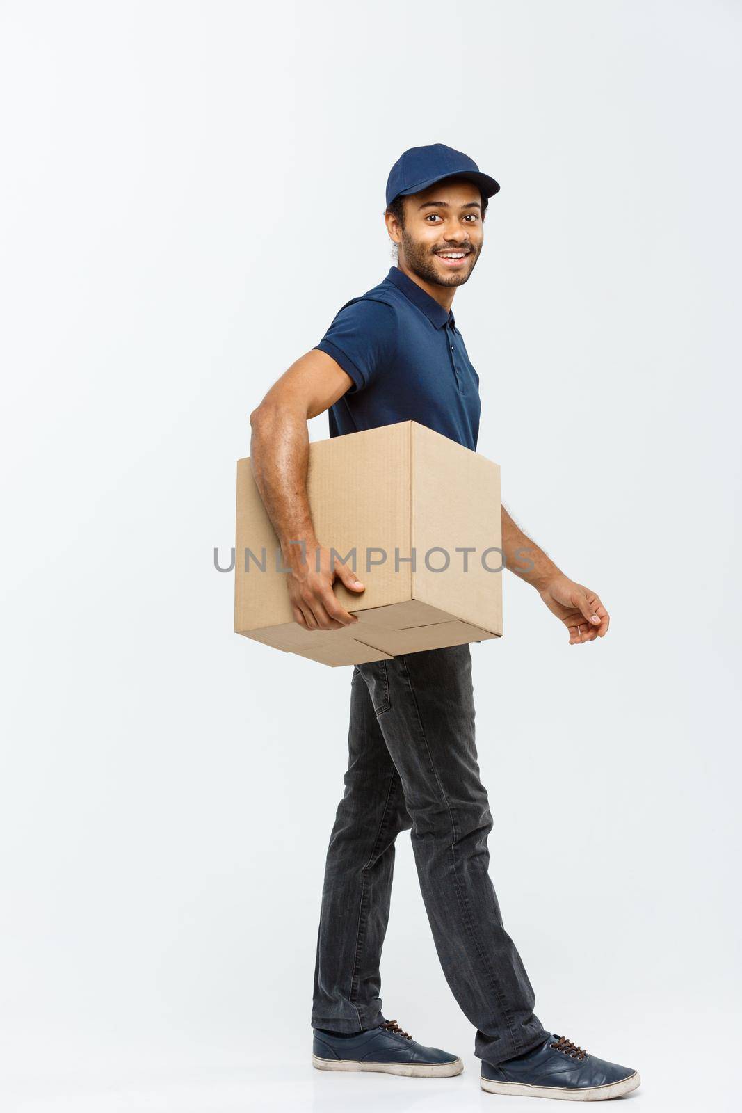 Delivery Concept - Portrait of Happy African American delivery man in blue cloth walking to send a box package to customer. Isolated on Grey studio Background. Copy Space