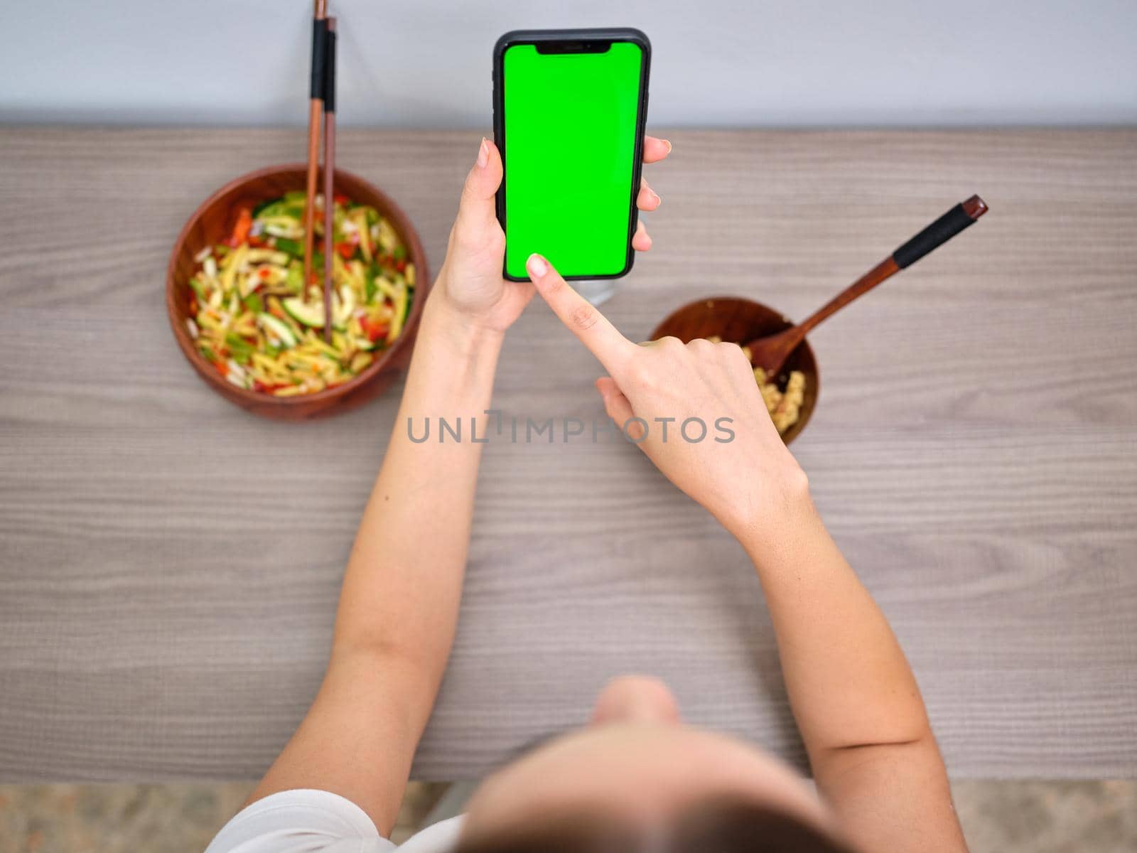 woman eating tabbouleh in a wooden bowl with chopsticks, looking at the green screen of her mobile phone, seen from above