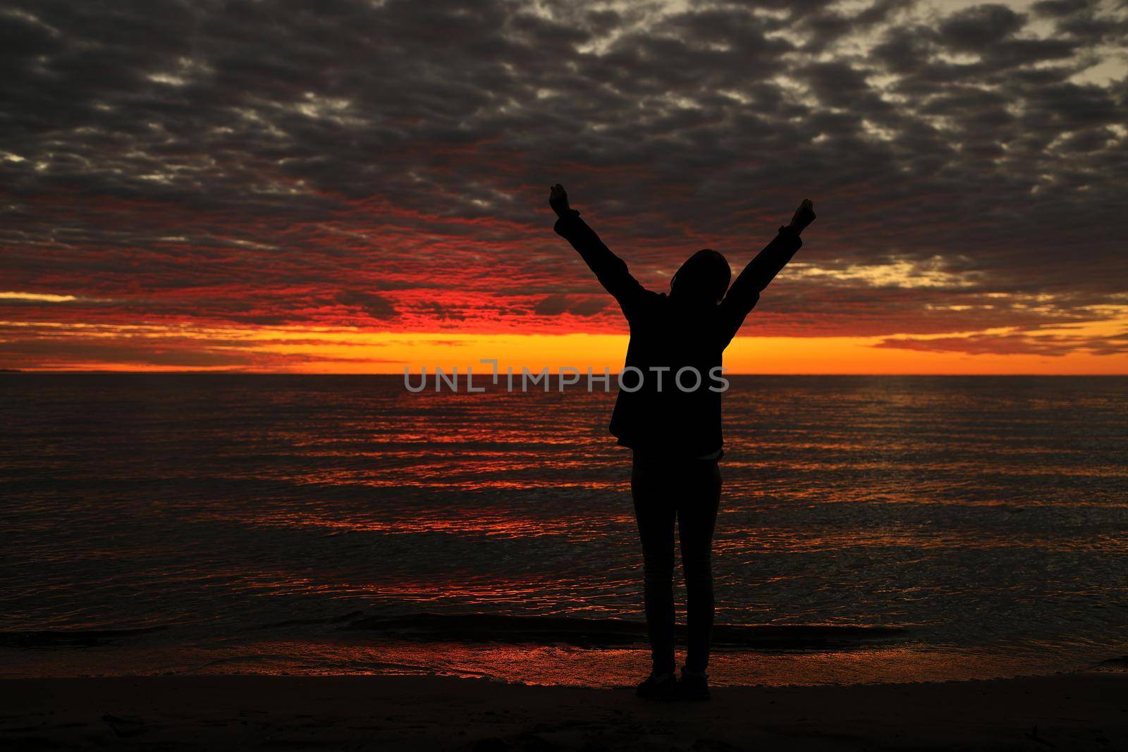 Happy woman sihouette with arms raised up in success to sunset glow at beach by water. Wellness, financial freedom, healthy life, happiness, success, victory concept background.