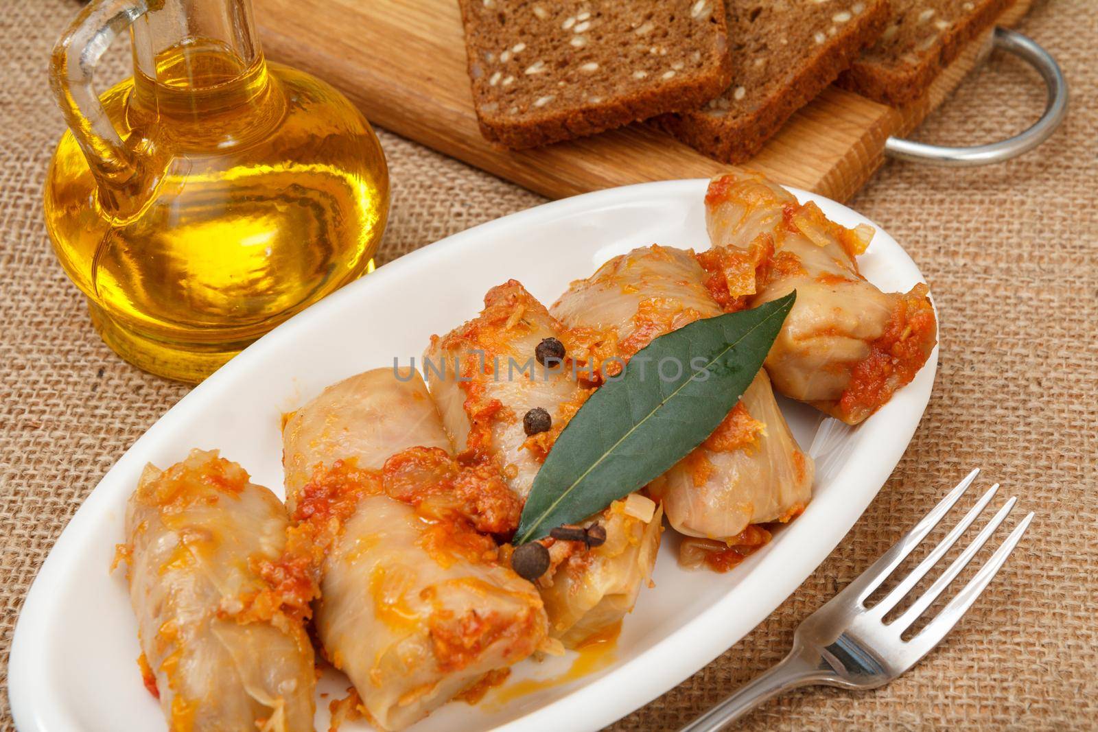 Delicious homemade cabbage rolls stuffed with rice and meat in white dish, fork, bottle with sunflower oil and bread on wooden cutting board on sackcloth