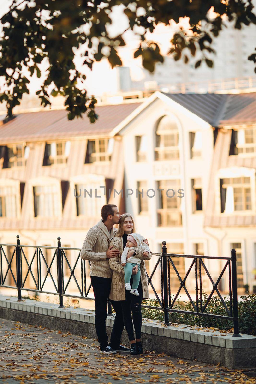 Side view of happy husband and wife with their lovely child. Handsome man holding little baby and beautiful woman hugging him from back. Young family in casual clothes walking together in sunny day.