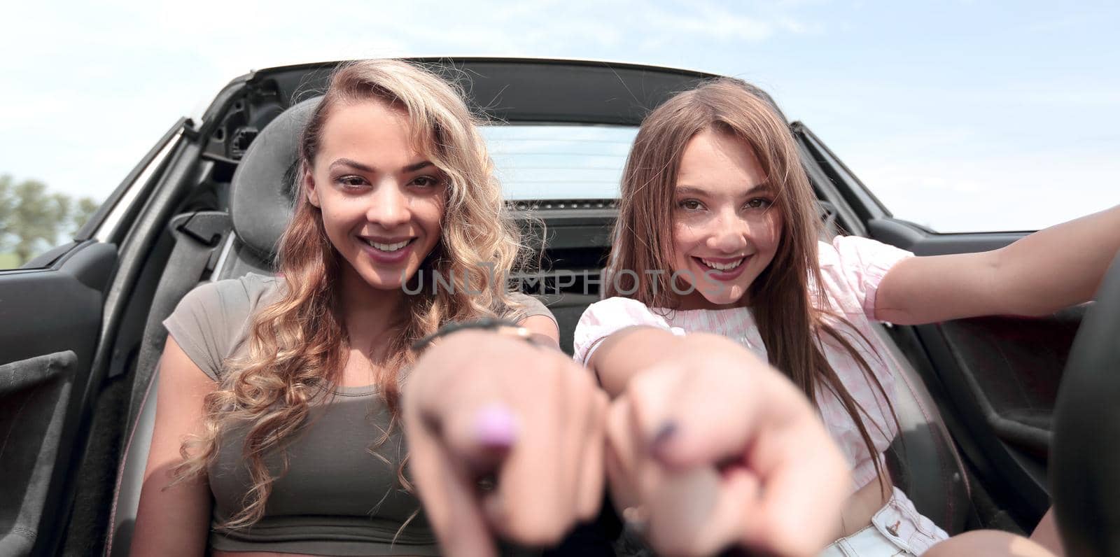 two happy girlfriends sitting in a car and pointing at you by asdf