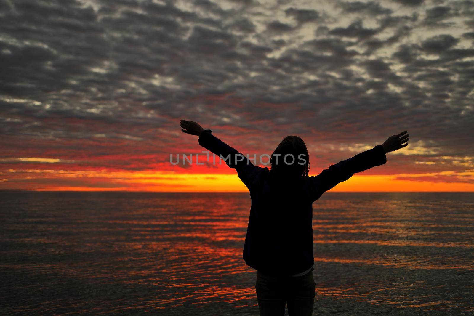 Happy woman sihouette with arms raised up in success to sunset glow at beach by water by markvandam