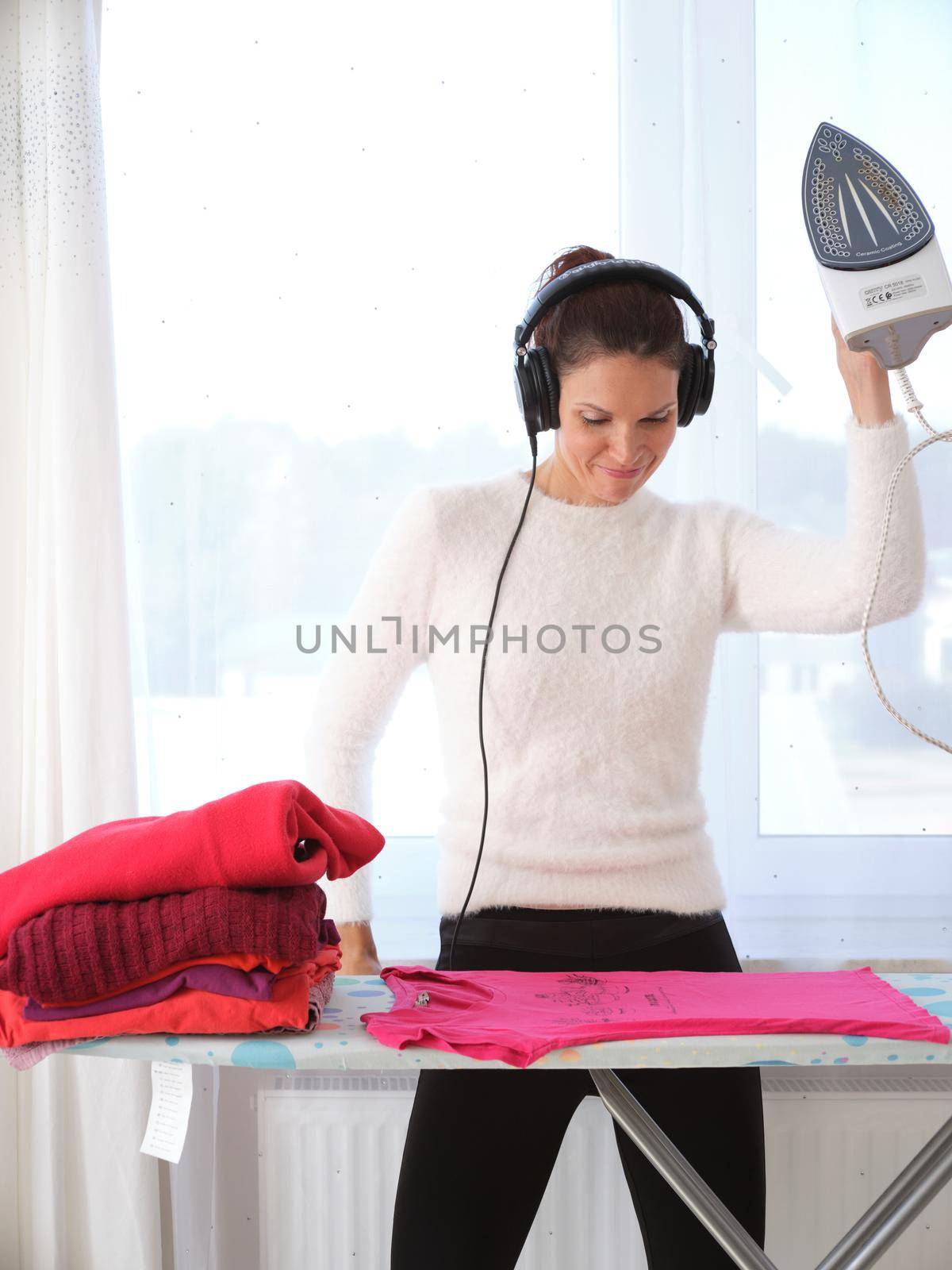 Vertical photo of a happy woman ironing while listening to music on her mobile phone at home