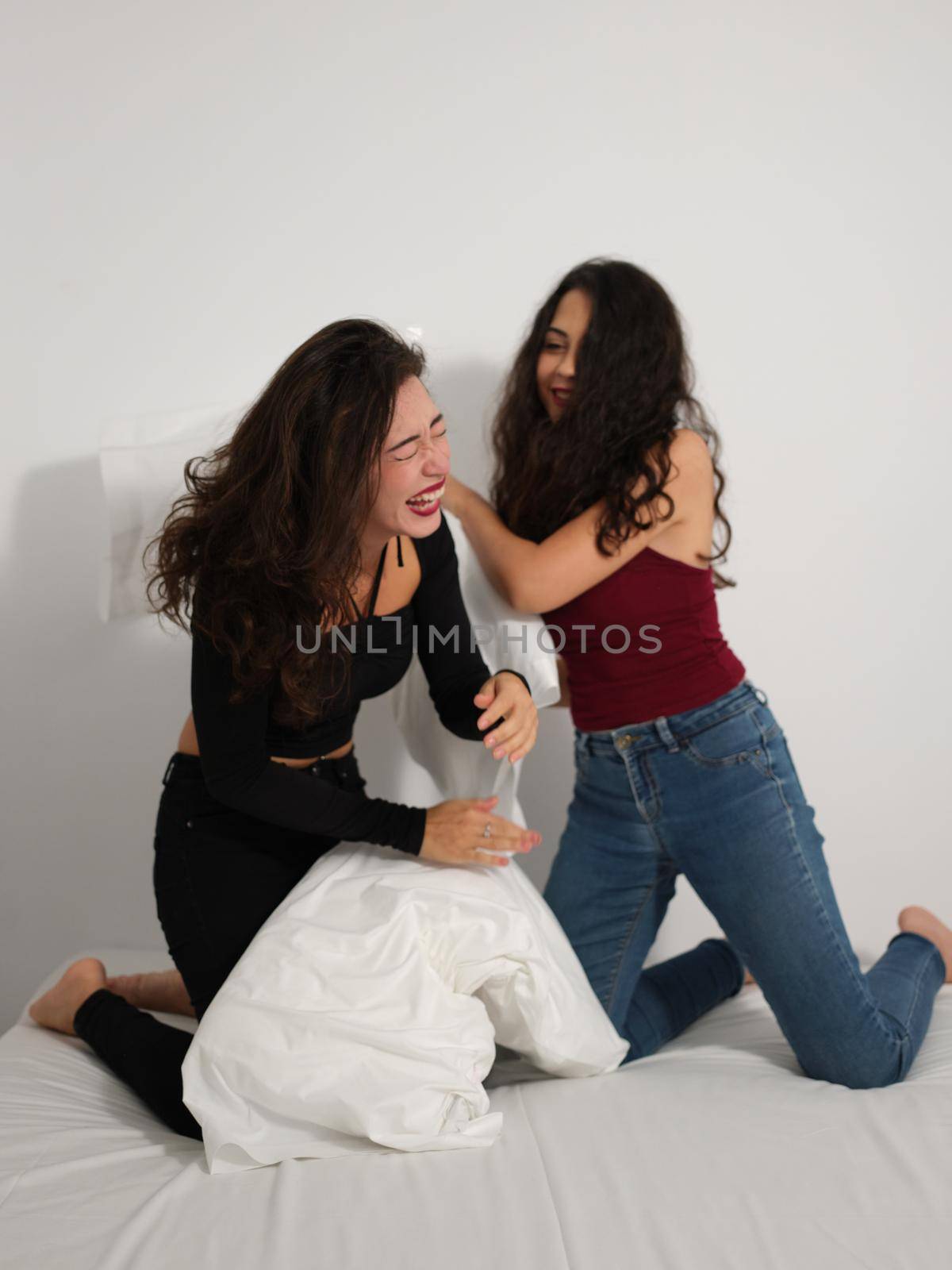 Vertical image of two friends kneeling on bed having fun and laughing playing pillow fight