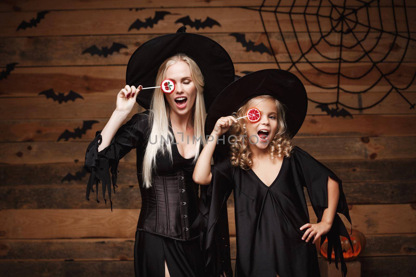 Halloween Concept: beautiful caucasian mother and her daughter in witch costumes celebrating Halloween with Halloween candy and sweet over bats and spider web on Wooden studio background