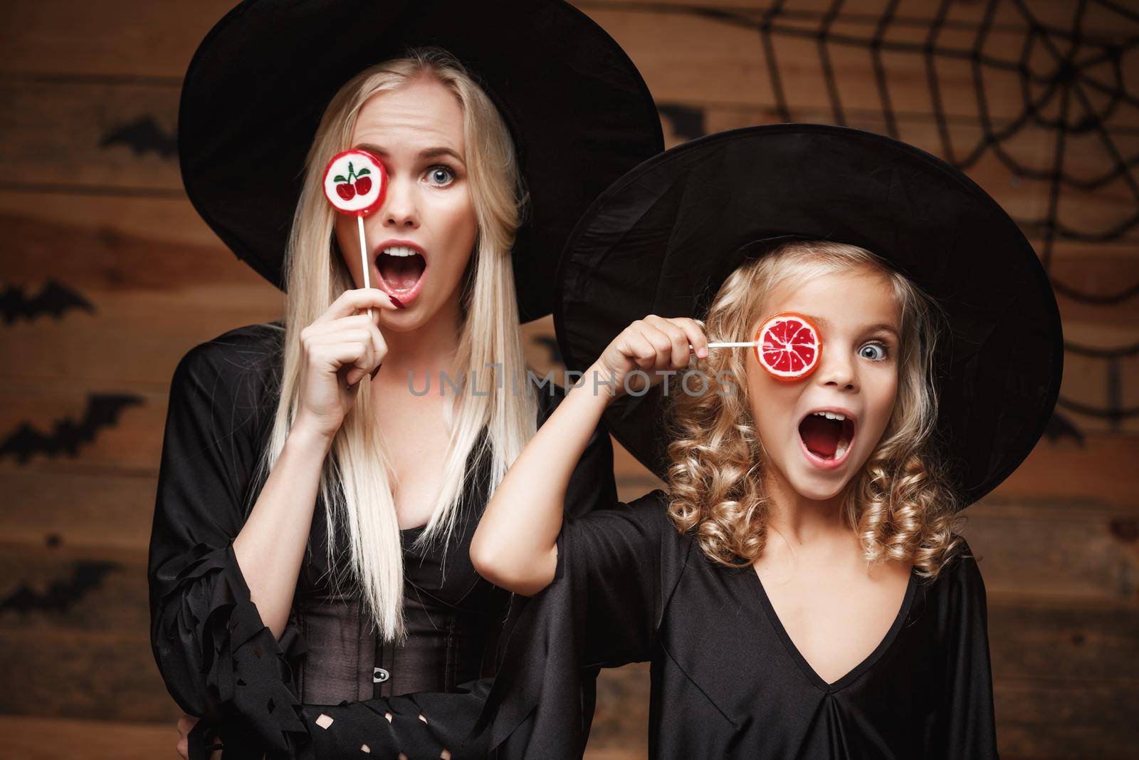 Halloween Concept - beautiful caucasian mother and her daughter in witch costumes celebrating Halloween with Halloween candy and sweet over bats and spider web on Wooden studio background. by Benzoix