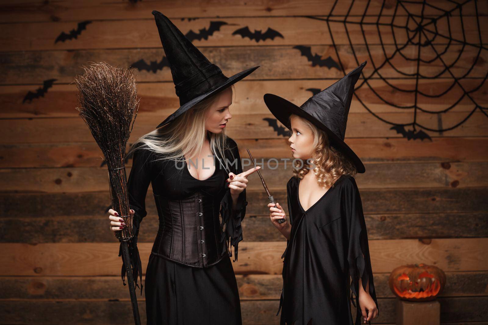 Halloween Concept - stressful witch mother teaching her daughter in witch costumes celebrating Halloween with curved pumpkins over bats and spider web on Wooden studio background. by Benzoix