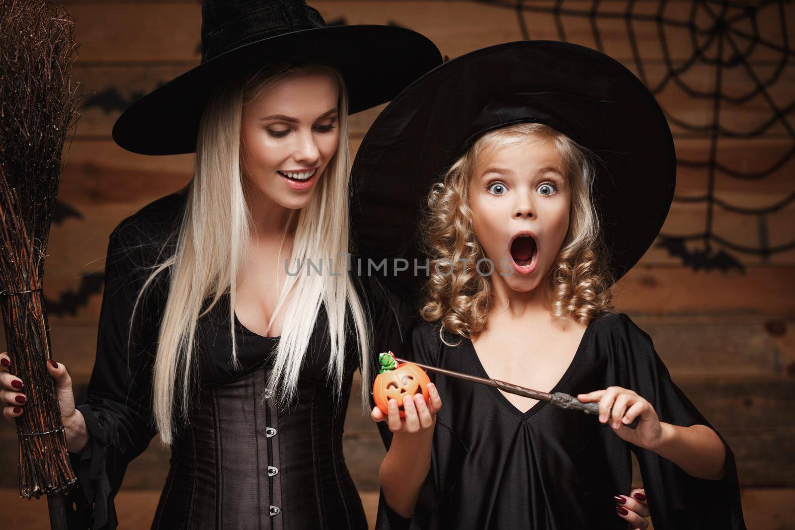 Halloween Concept: beautiful caucasian mother and her daughter in witch costumes enjoy using magic with magic wand to halloween pumpkin jar over bats and spider web on Wooden studio background.