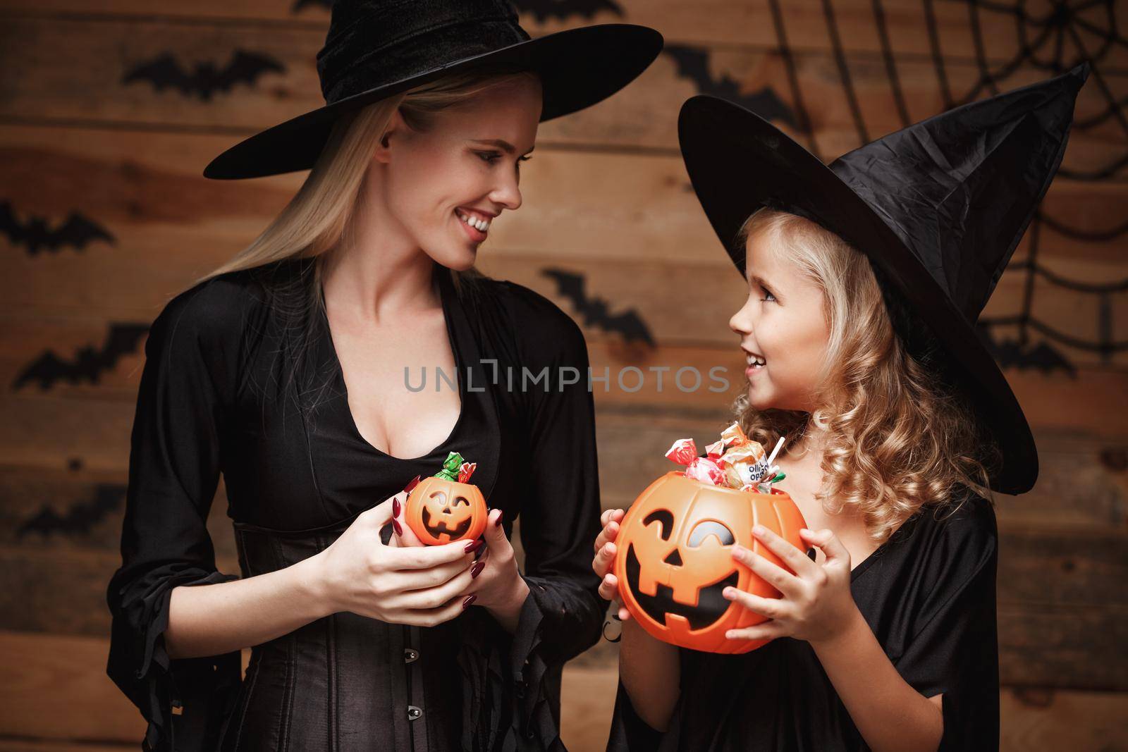 Halloween Concept - beautiful caucasian mother with disappointed feeling with happy little daugther enjoy with Halloween candy and sweet over bats and spider web on Wooden studio background. by Benzoix