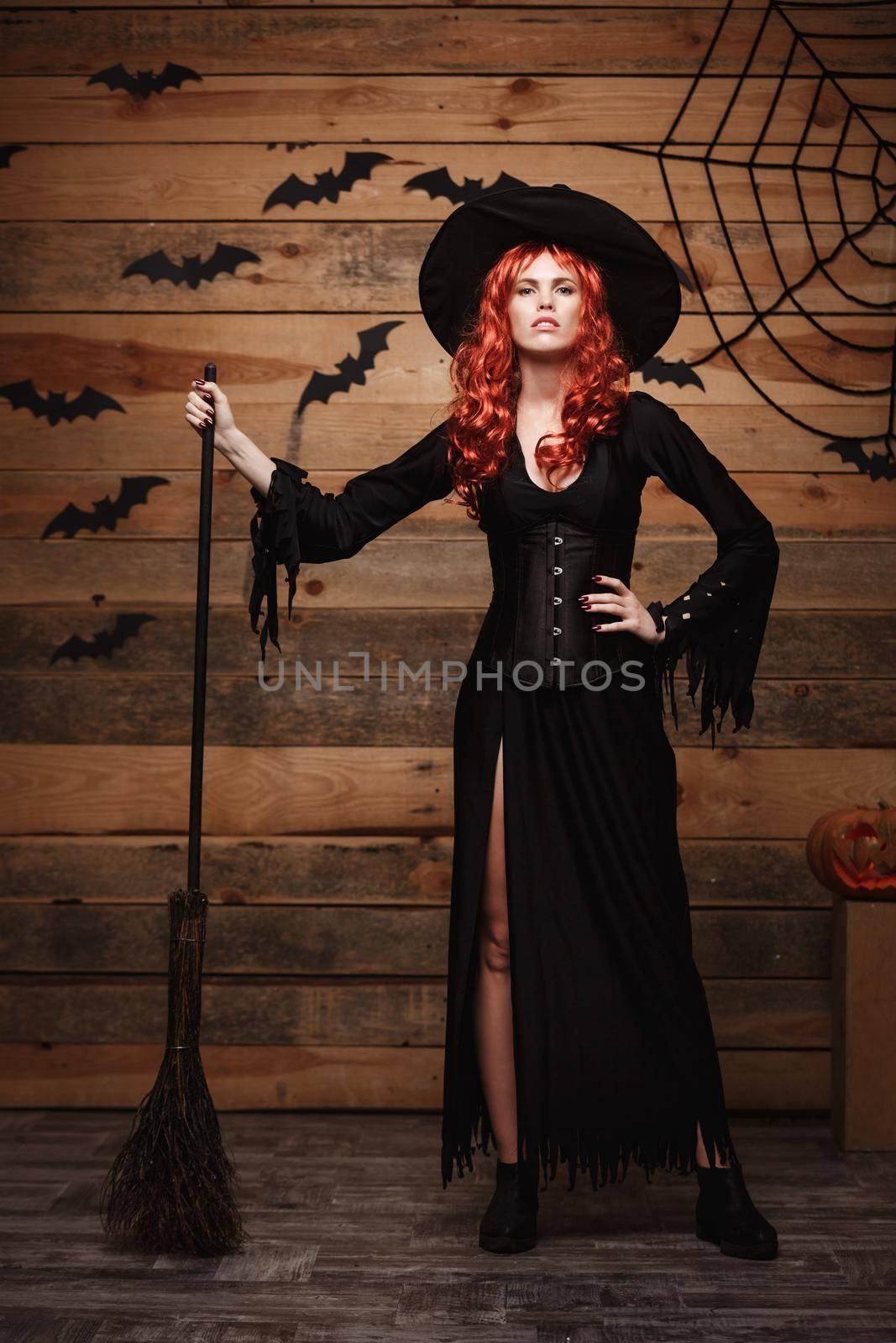 Halloween witch concept - Full-length Happy Halloween red hair Witch holding posing with magic broomstick over old wooden studio background. by Benzoix