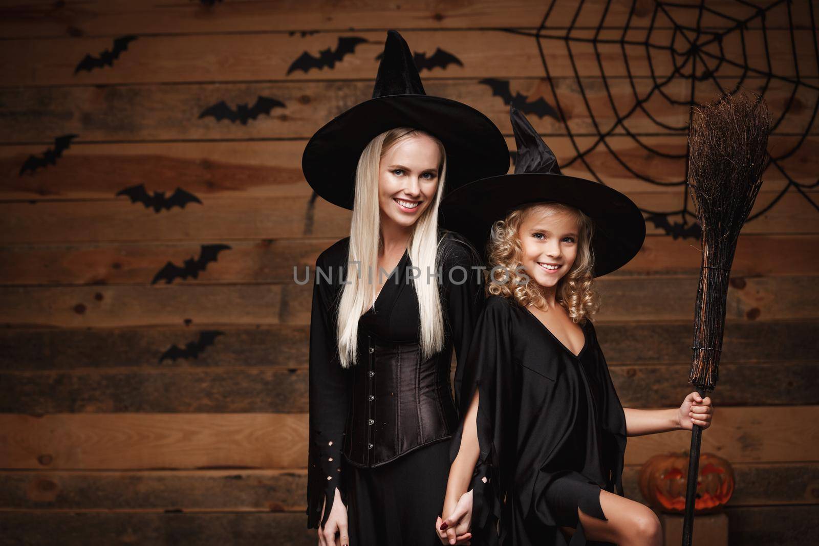 Halloween Concept - cheerful mother and her daughter in witch costumes celebrating Halloween posing with curved pumpkins over bats and spider web on Wooden studio background